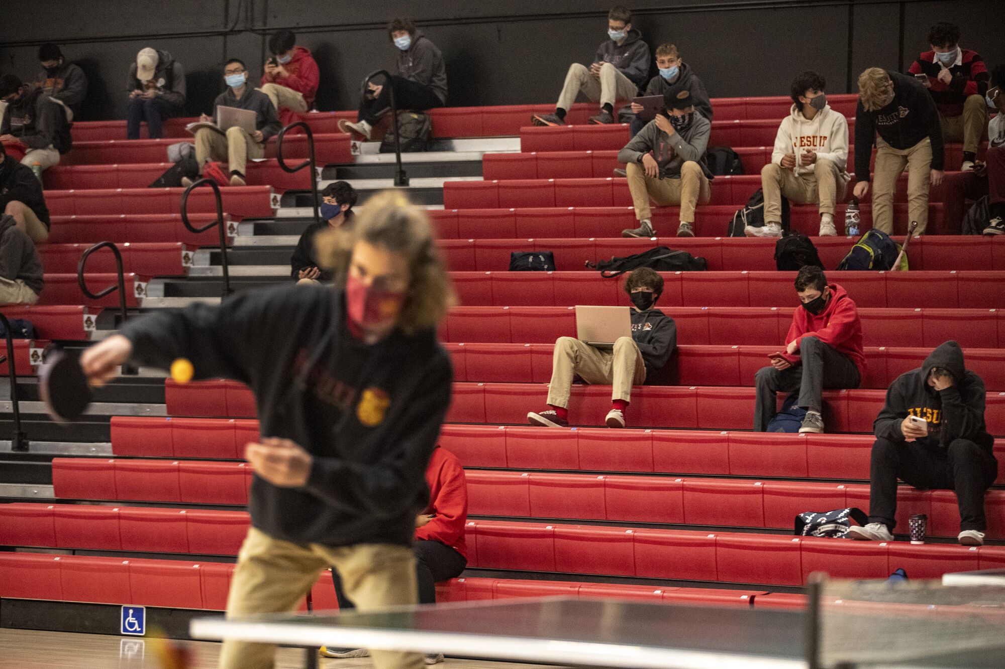 A student plays pingpong while others sit in the school gymnasium bleachers.
