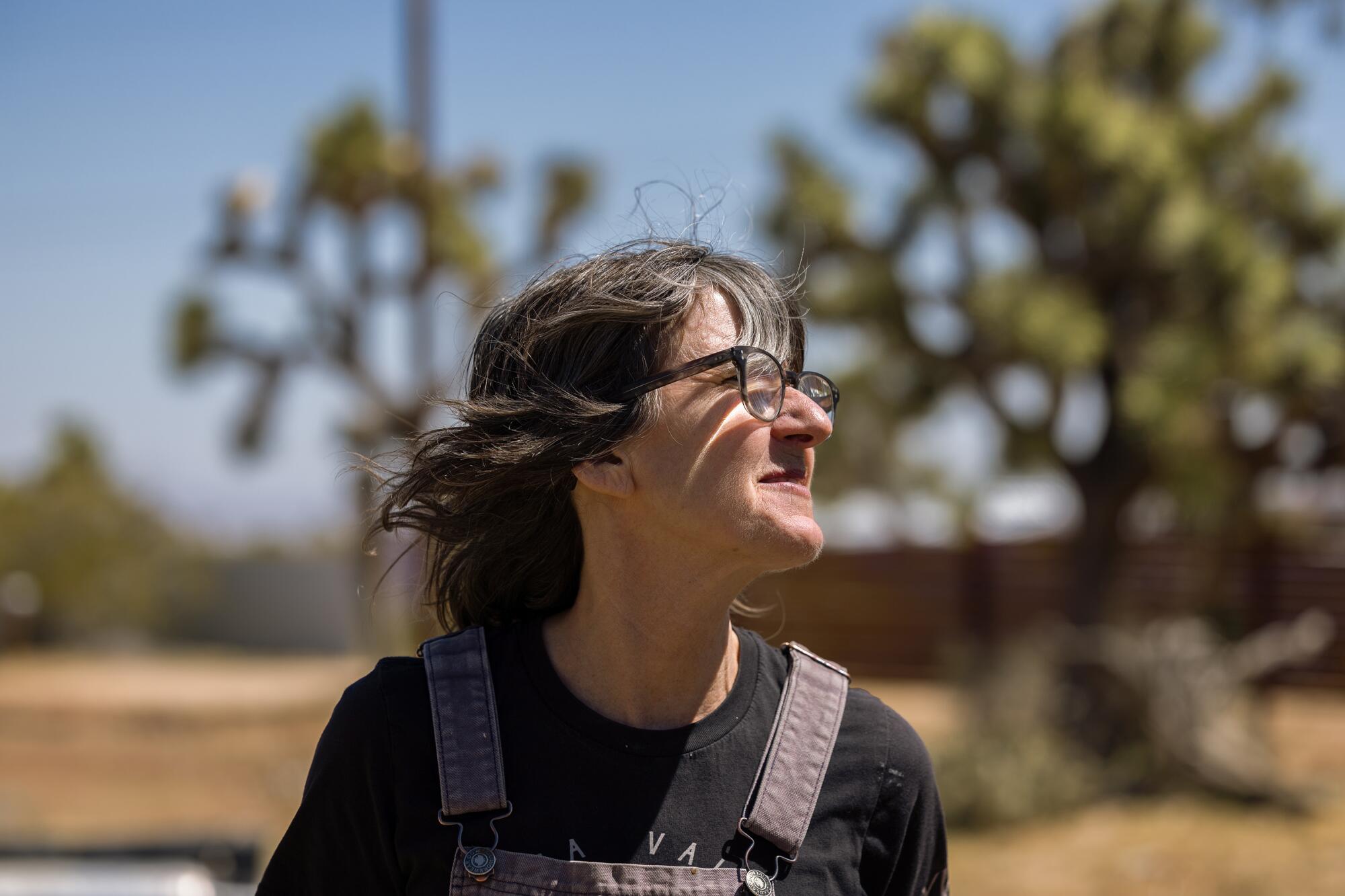 Heidi Schwegler, wearing glasses and overalls, looks right as she poses in front of Joshua trees.