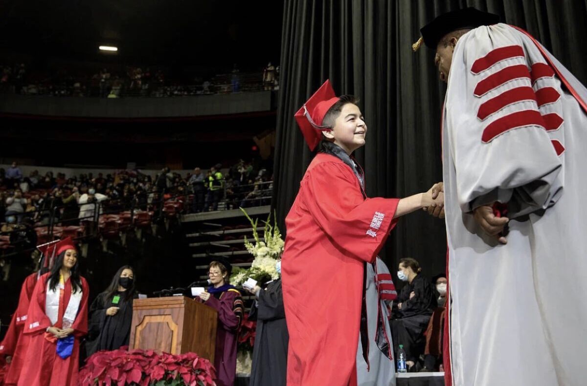 A boy wearing graduation robes shakes the hand of a faculty member on stage