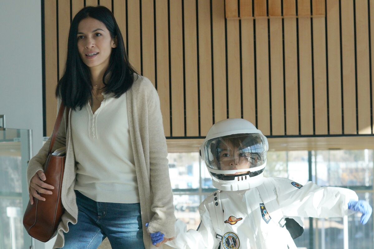 A woman holds the hand of a child in a spacesuit.