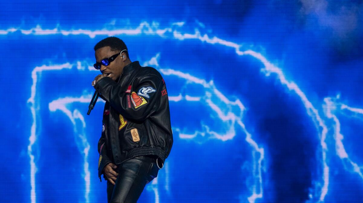 A man in sunglasses and all black leather raps into a microphone in front of a dramatically lit background