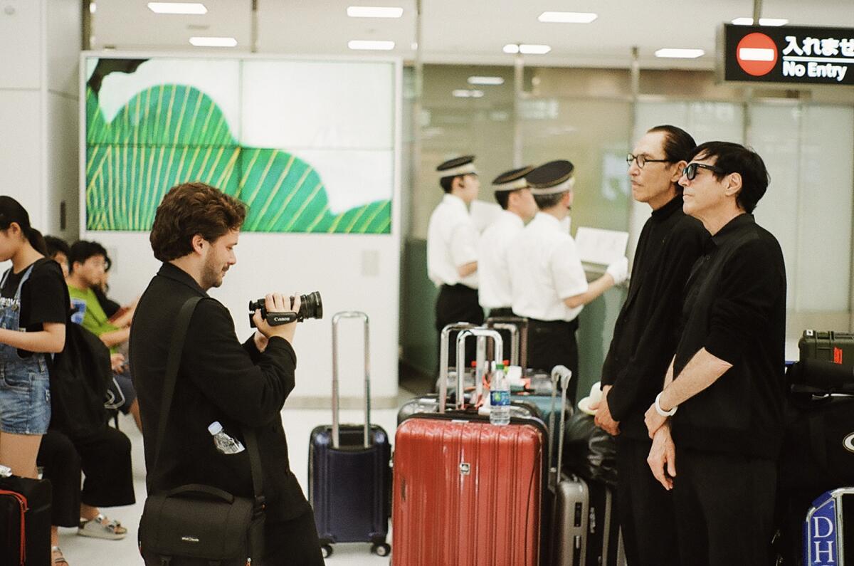A man at left holds up a camera facing two men standing together at right. Luggage is in the background. 