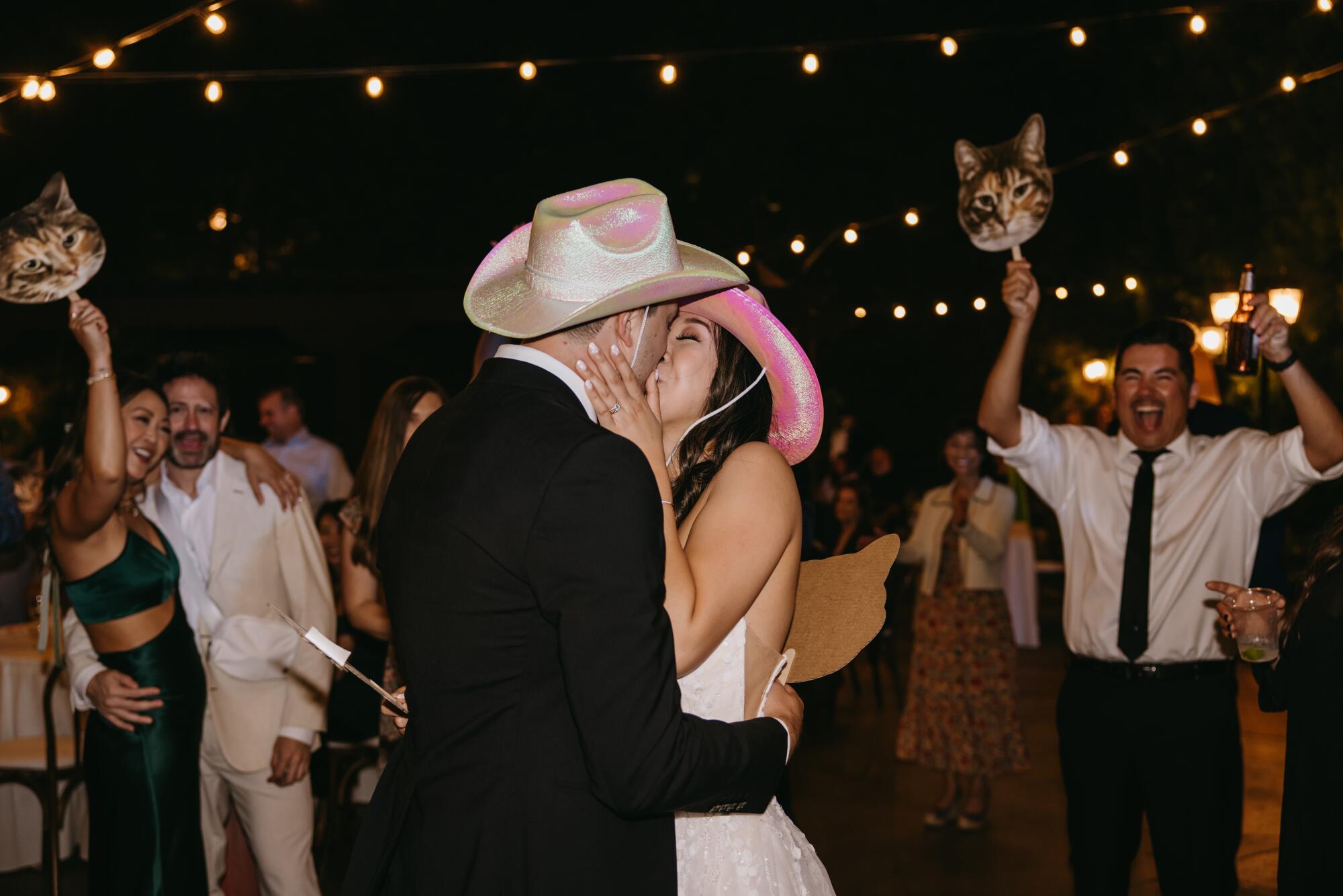 A couple wearing cowboy hats and kissing as people cheer behind them