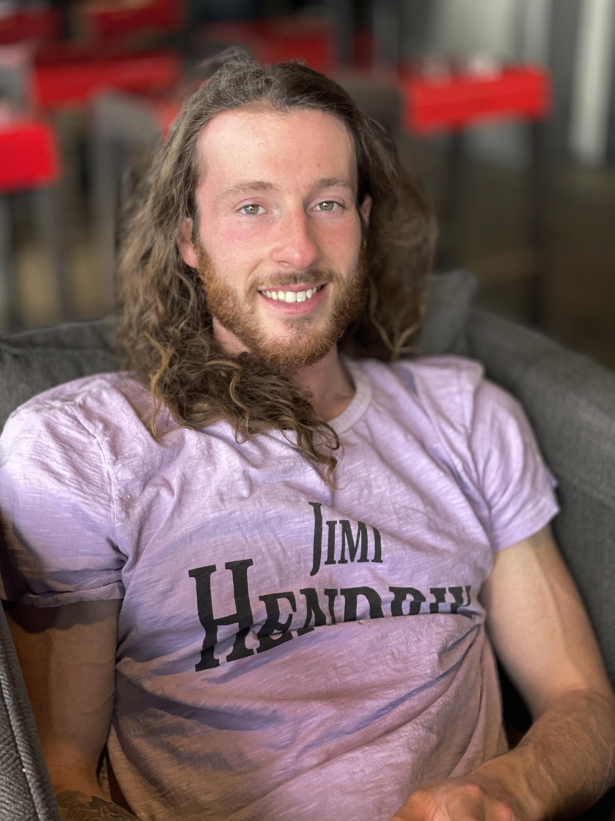 A bearded man with long, wavy brown hair and wearing a pale pink T-shirt sits smiling