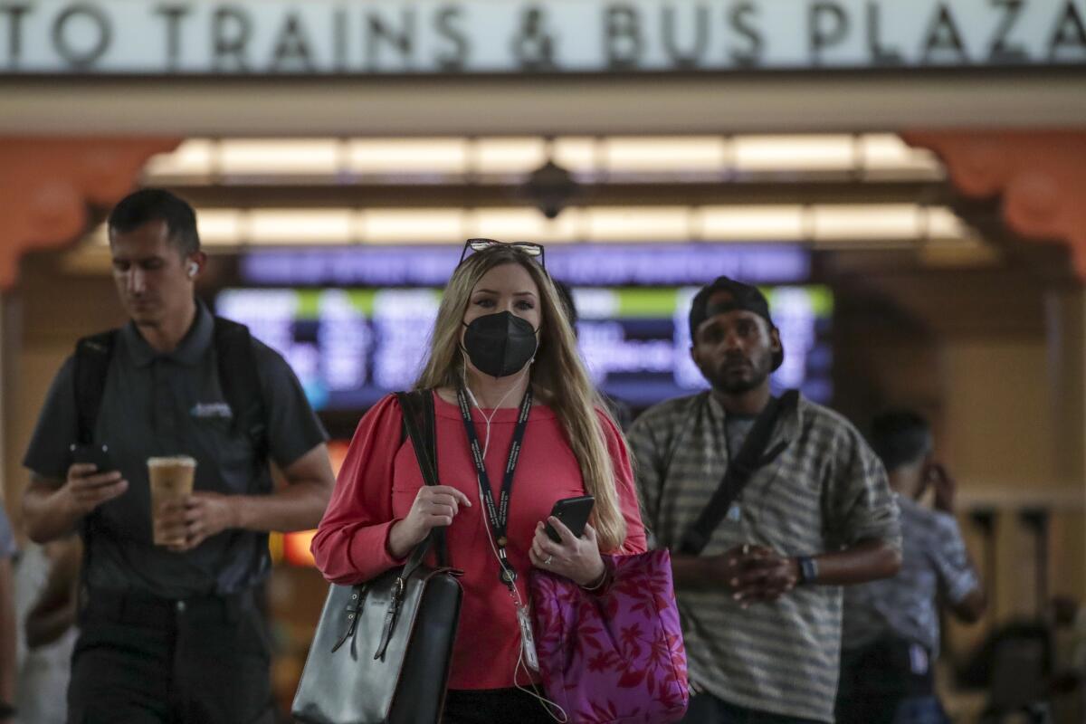 A woman in a mask walks in front of two men without masks in a train station
