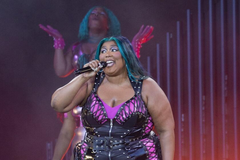 Lizzo sings into a microphone while wearing a black leather outfit over a pink bodysuit. She has blue streaks in her hair.