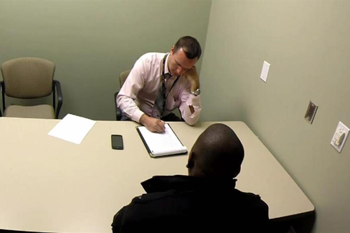 A detective taking notes sits across a table from another person.