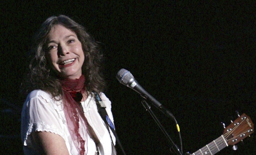 A smiling woman in a white shirt and red scarf holds a guitar as she stands at a microphone.
