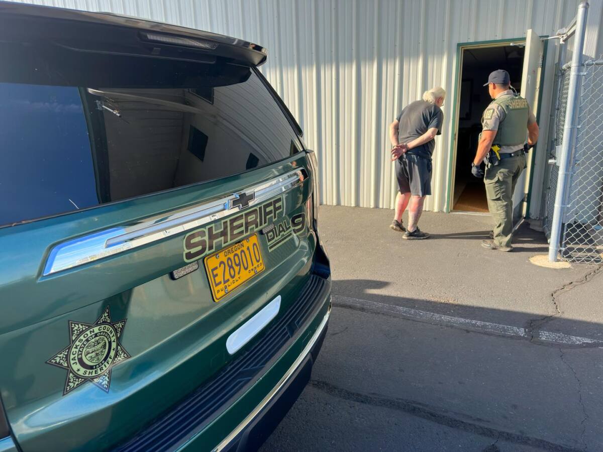 A sheriff's deputy holds open a door for an older man in handcuffs.