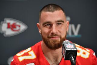 NFL star Travis Kelce speaks at a microphone while seated in his Kansas City Chiefs uniform