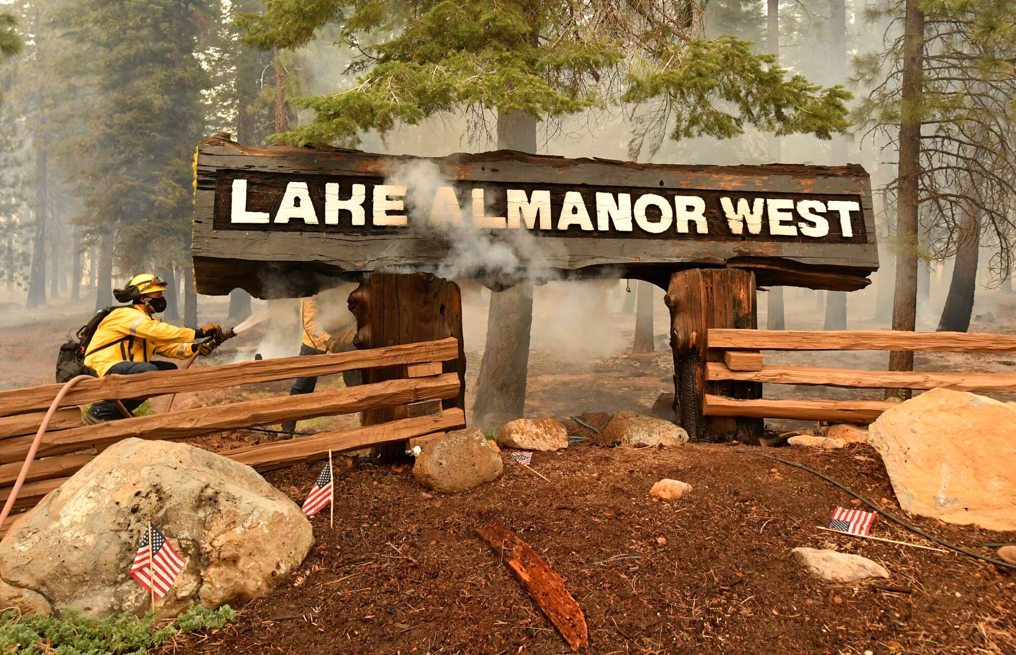 Two firefighters aim their hoses at a smoking wooden sign that says "Lake Almanor West."