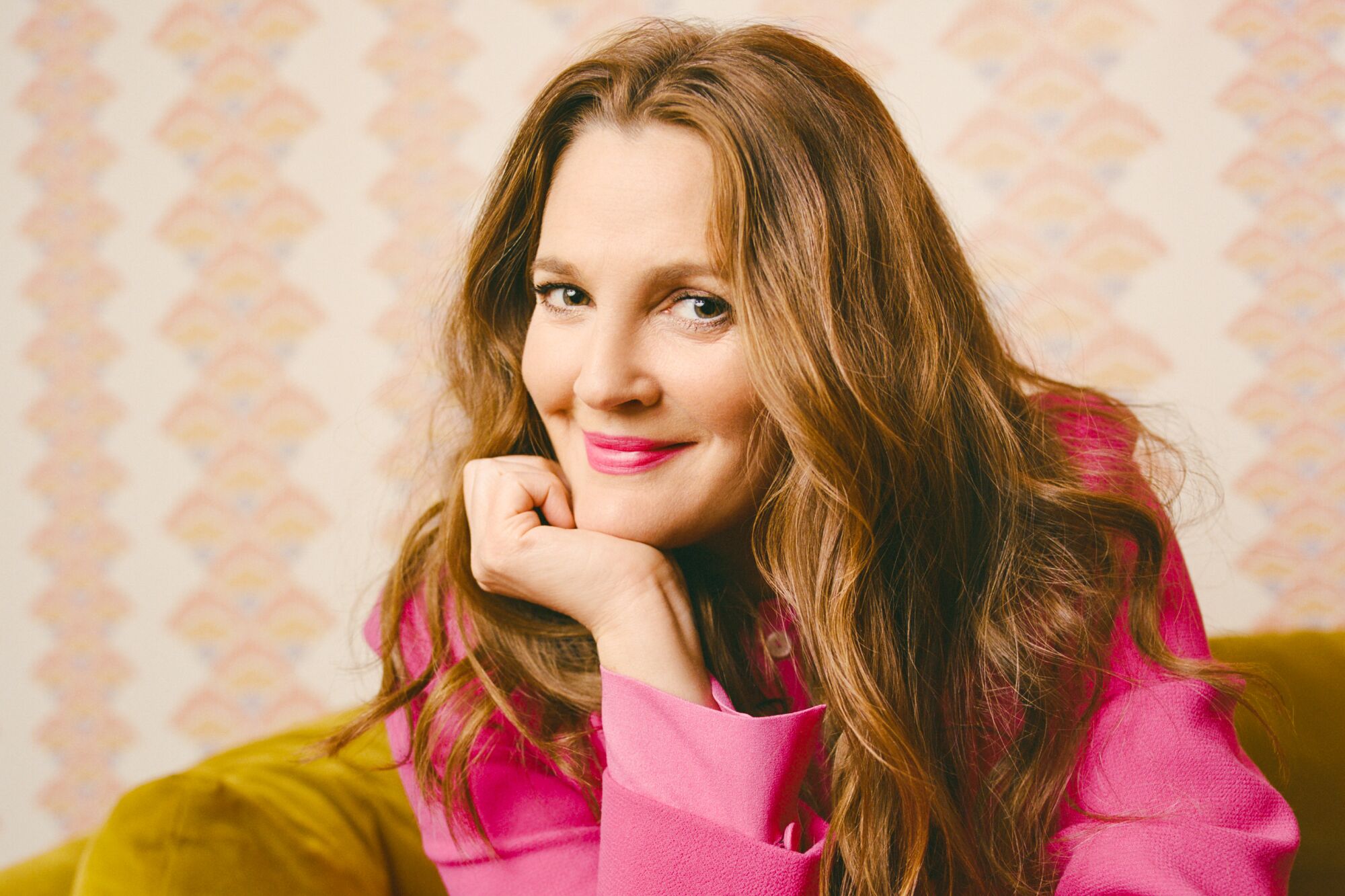 Drew Barrymore, wearing a pink shirt, poses with her hand under her chin.