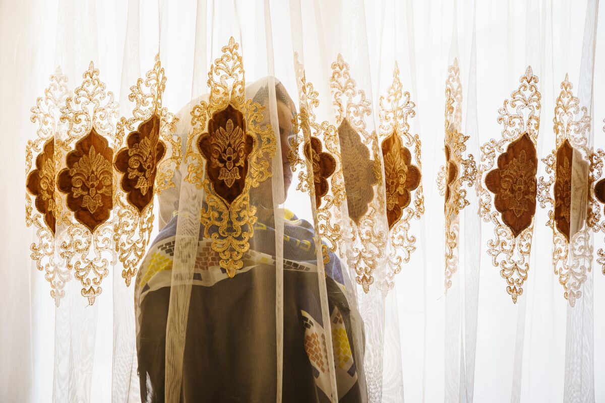 A woman in a head scarf stands behind a sheer curtain, obscuring her features. 