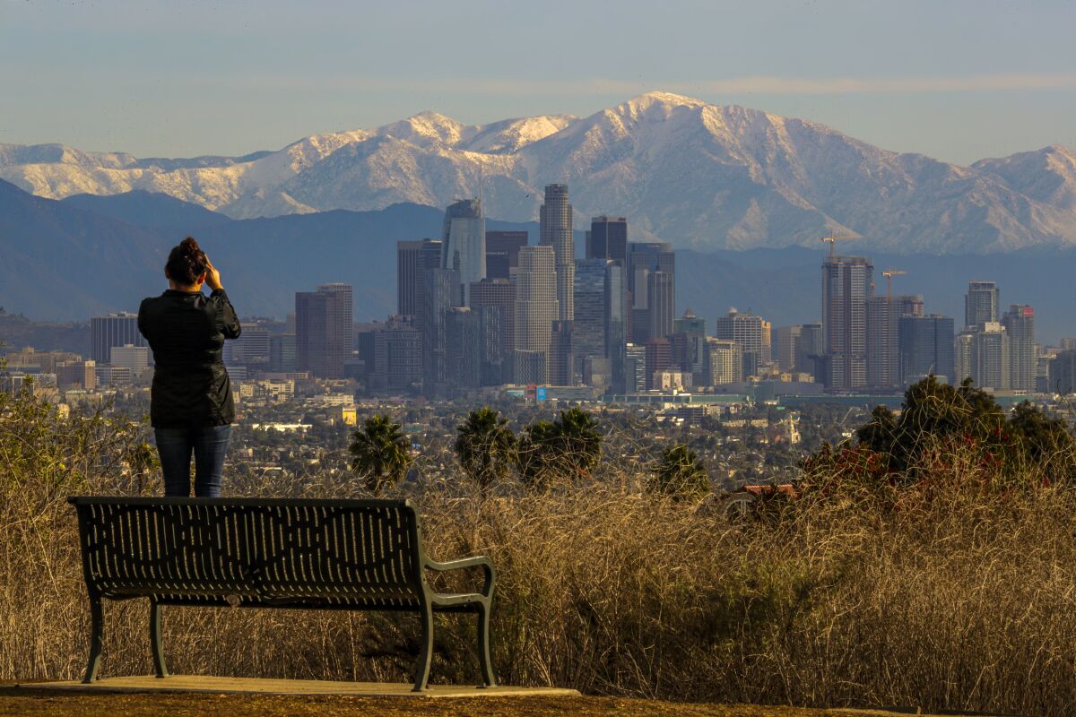 A person stands on a park bench taking a photo of an urban skyline backed by snowy mountains.