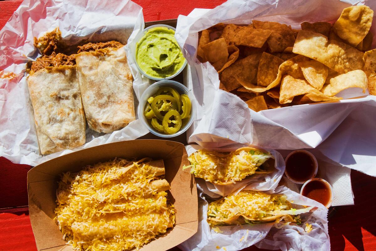 An overhead photo of a cardboard box filled with rolled corn burritos, a chile colorado burrito, hard tacos and flour chips.