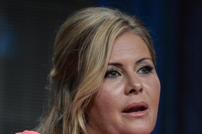 Nicole Eggert wears a pink dress and a gold necklace as she speaks while seated at an event