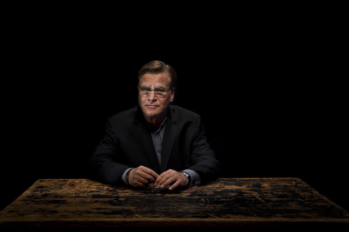 A man in glasses sits at a desk in a darkened room.