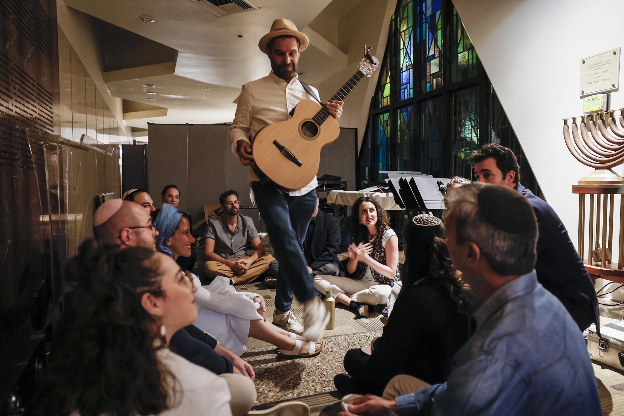 A man standing and holding a guitar, surrounded by several people seated on the floor.
