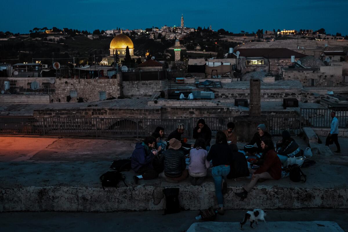 A group of young people sit and eat on a rooftop with a view of buildings, one with a bright golden dome, as darkness falls
