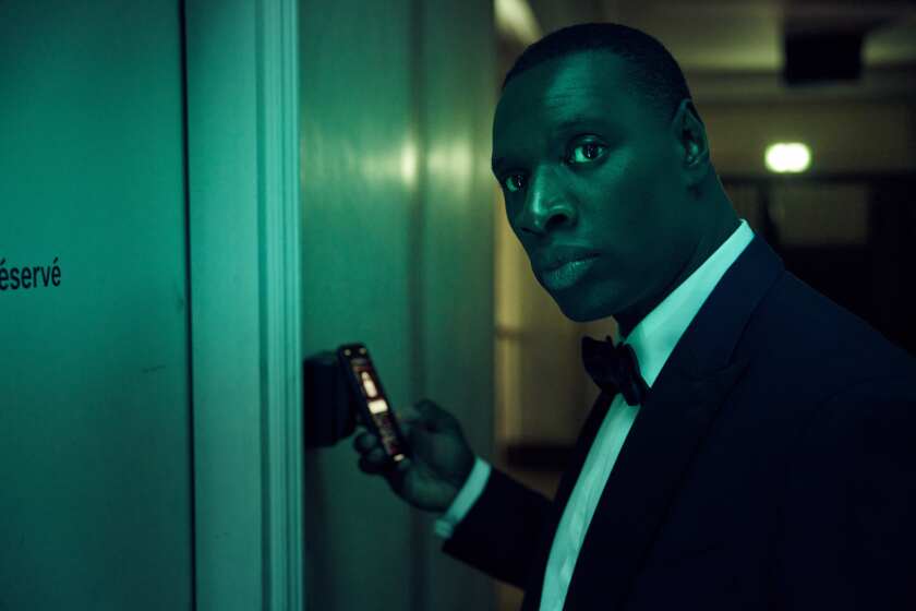A man in a tuxedo holding a cellphone looks down a corridor.