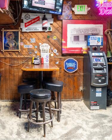 A table with bar stools and an ATM on a sawdusty floor next to a wood-paneled wall in a dive bar