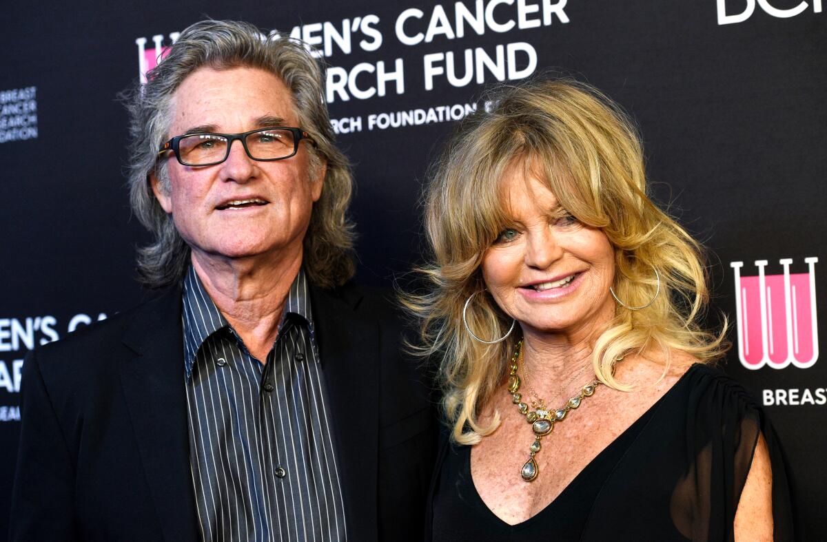Kurt Russell wearing a black jacket and dark striped shirt standing next to Goldie Hawn, who is in a black V-neck dress