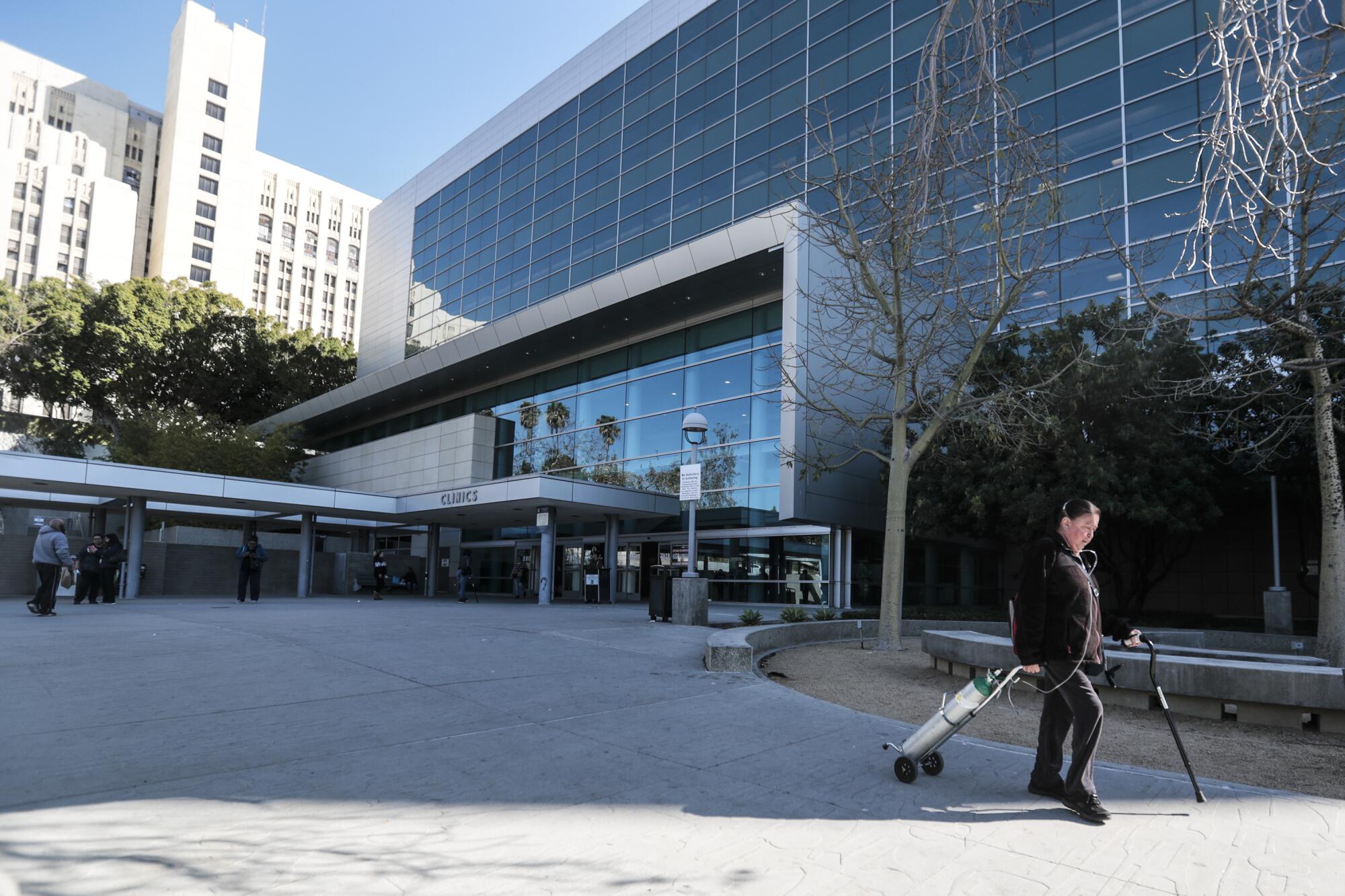 A person with a cane and oxygen tank walks outside a hospital building