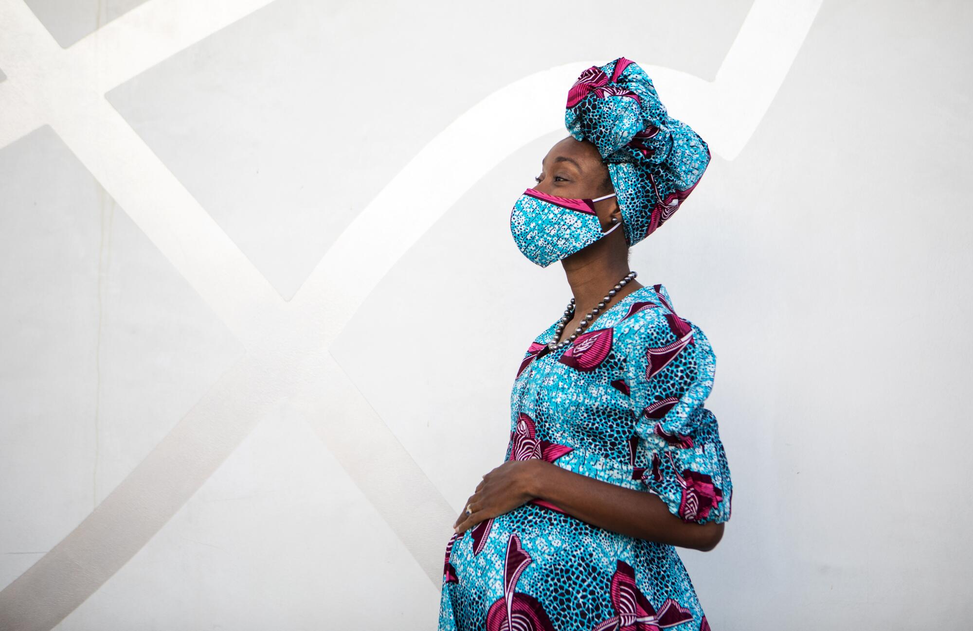 A pregnant Black woman wearing a matching turquoise dress, headscarf and face mask rests her hand on her belly
