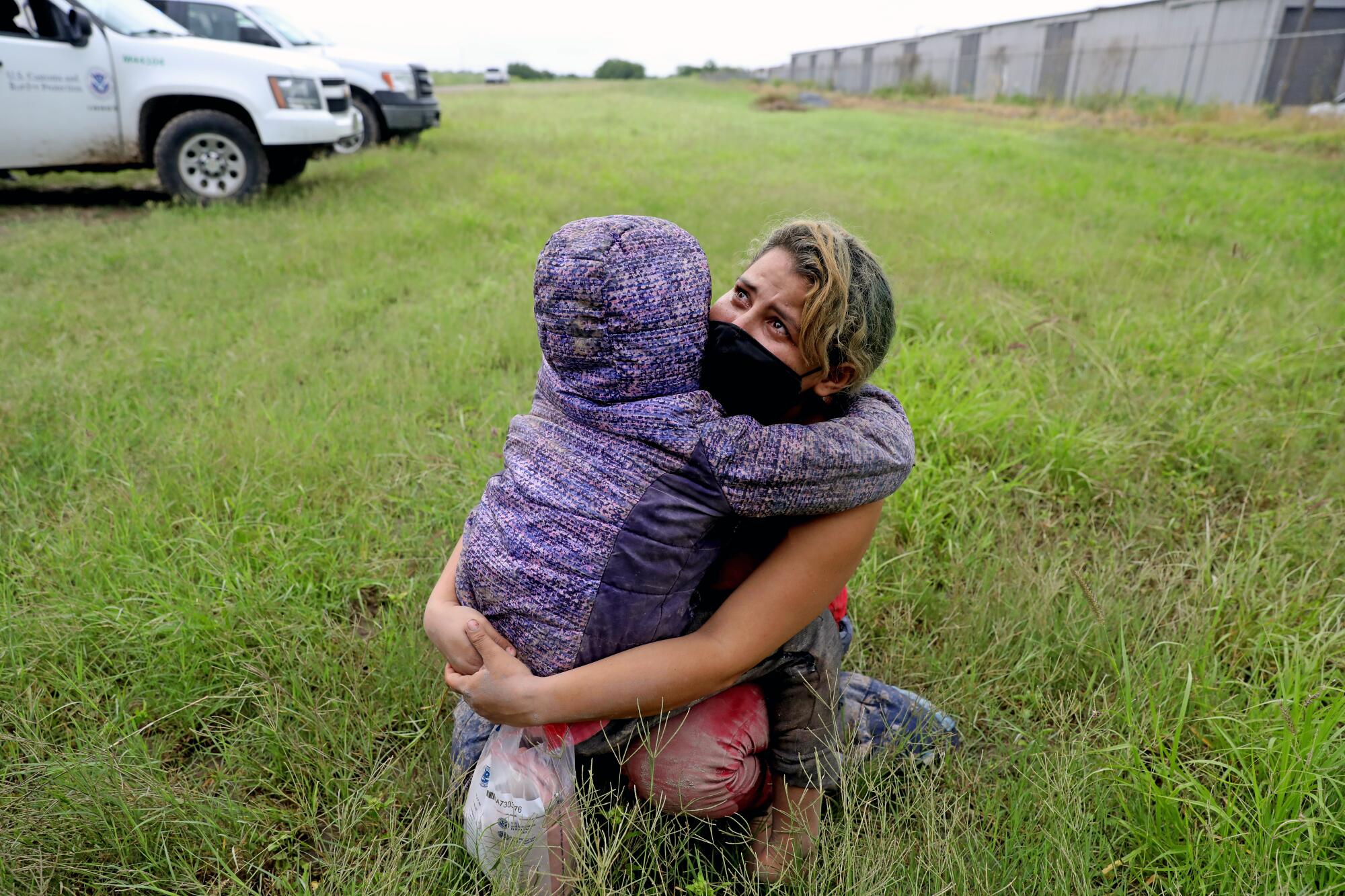 A woman hugs a child, left, in a purple hooded jacket on grassy area
