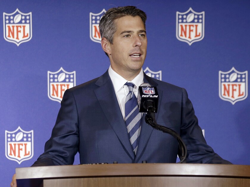 A man in a suit standing at a lectern in front of a backdrop with NFL logos
