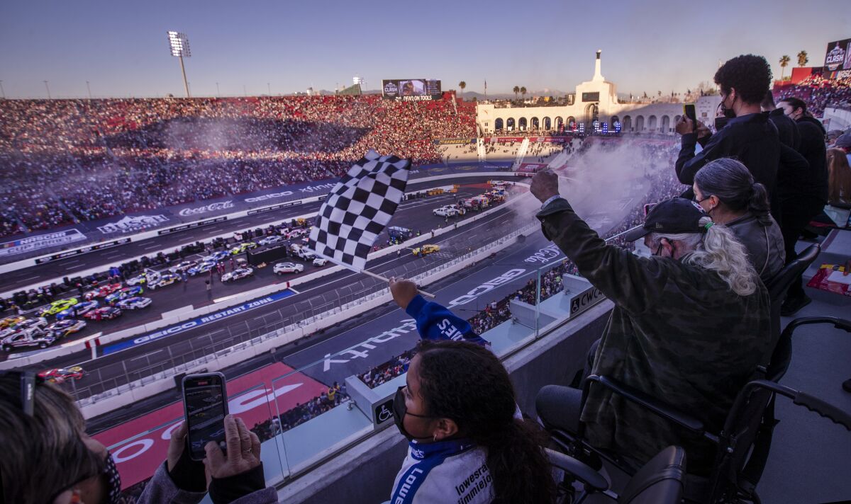 A group of people wear masks and wave flags as they look down at a race car track from stadium seats
