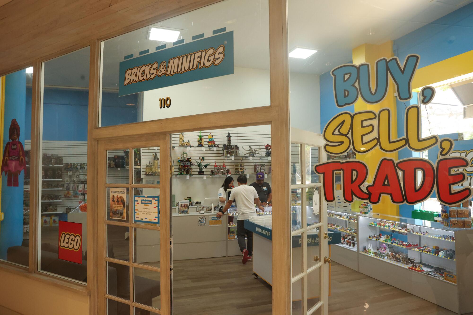 A storefront with windowed doors and windows labeled "Bricks & Minifigs" and "Buy, Sell, Trade"