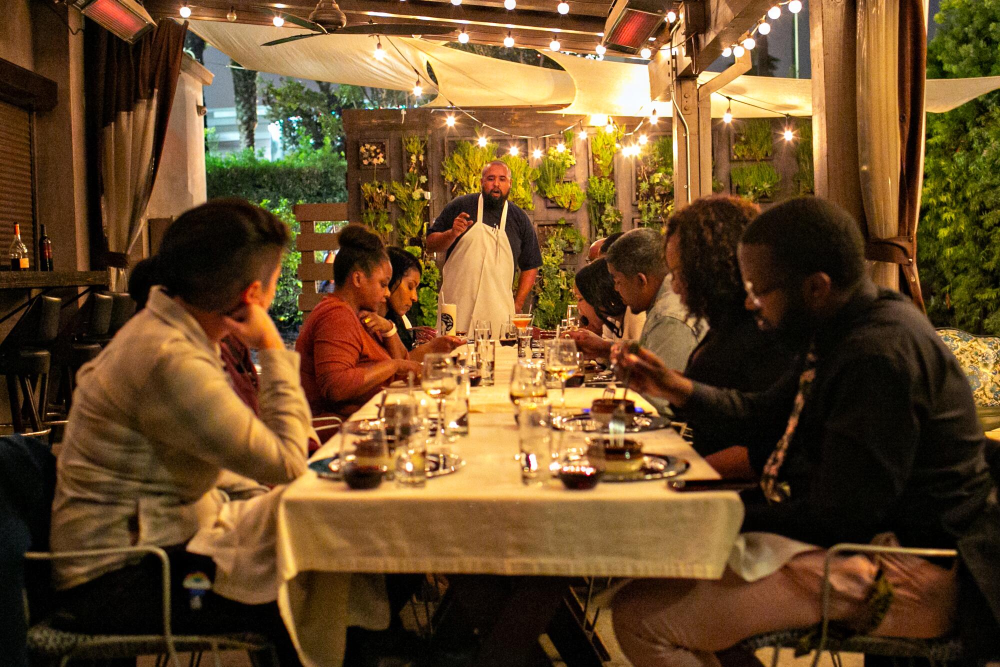 A chef stands before seated guests at a table, discussing a meal.
