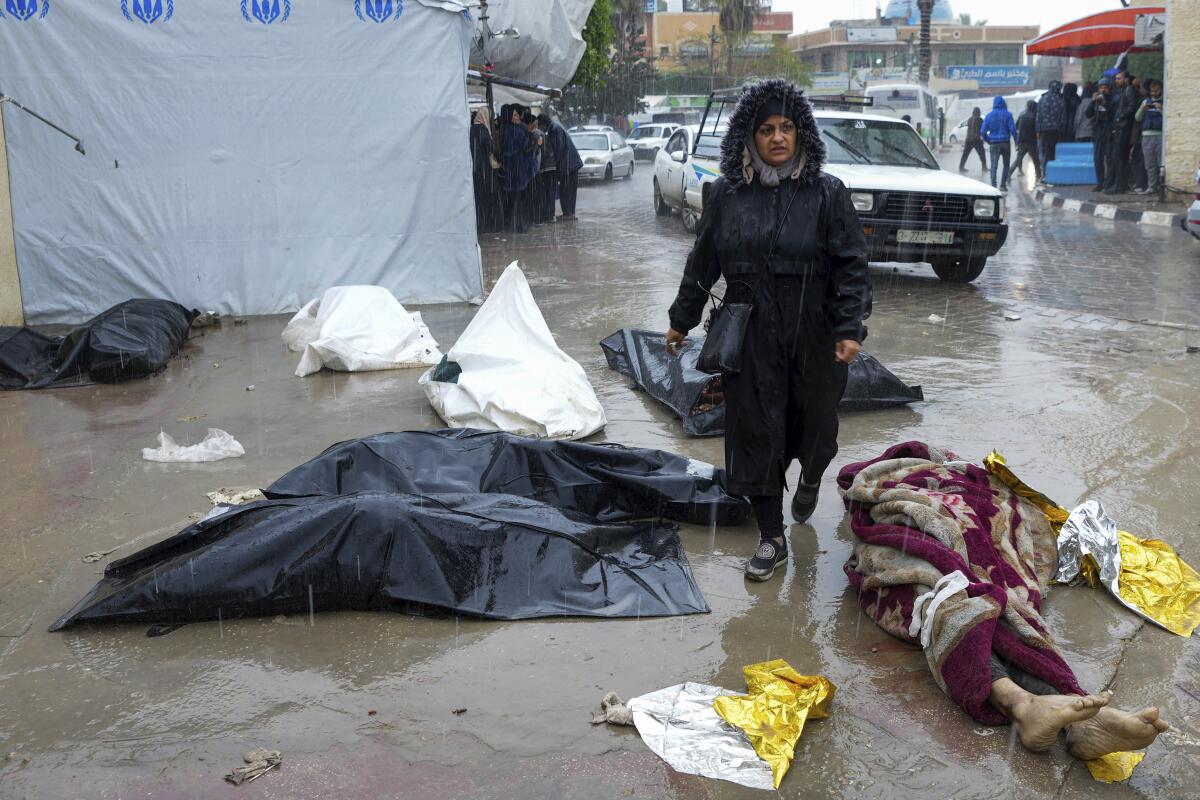 A woman walks between bodies covered in blankets or on bags in front of a morgue in Deir al Balah, Gaza Strip.