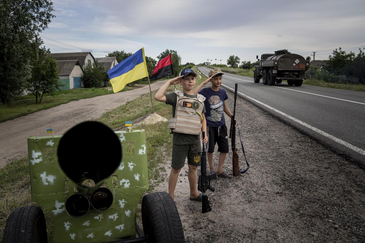 Two boys standing in front of a pair of flags beside a road salute while holding plastic guns