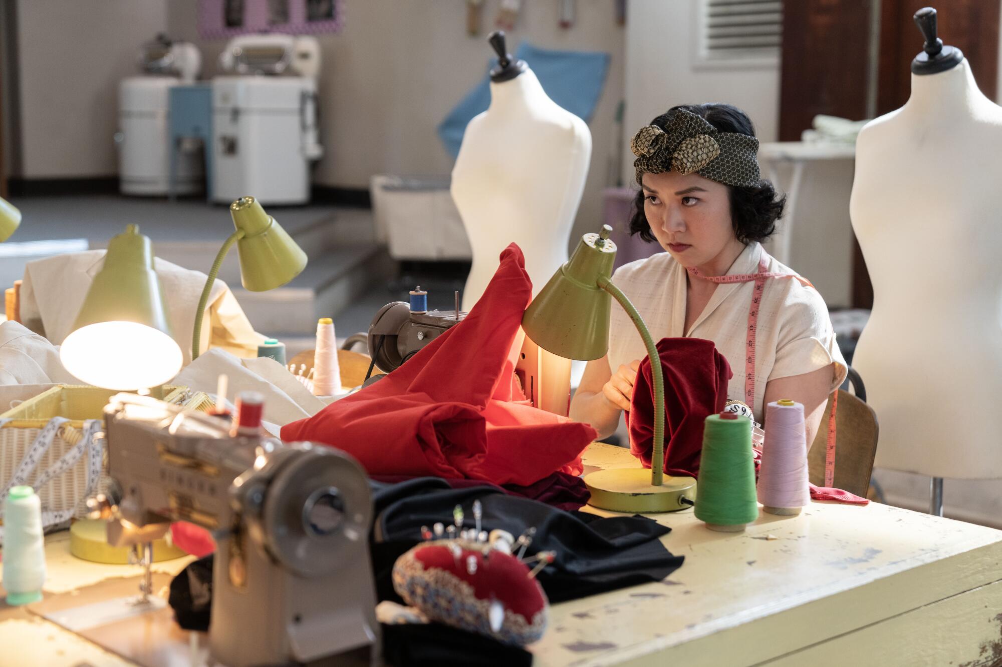 A young woman sits among fabric, thread and a sewing machine in a scene from "Grease: Rise of the Pink Ladies."