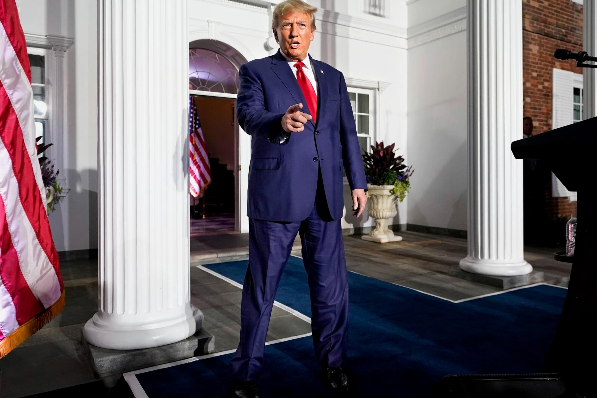 A full-body frame of Donald Trump standing at the center between two white columns pointing toward the camera 