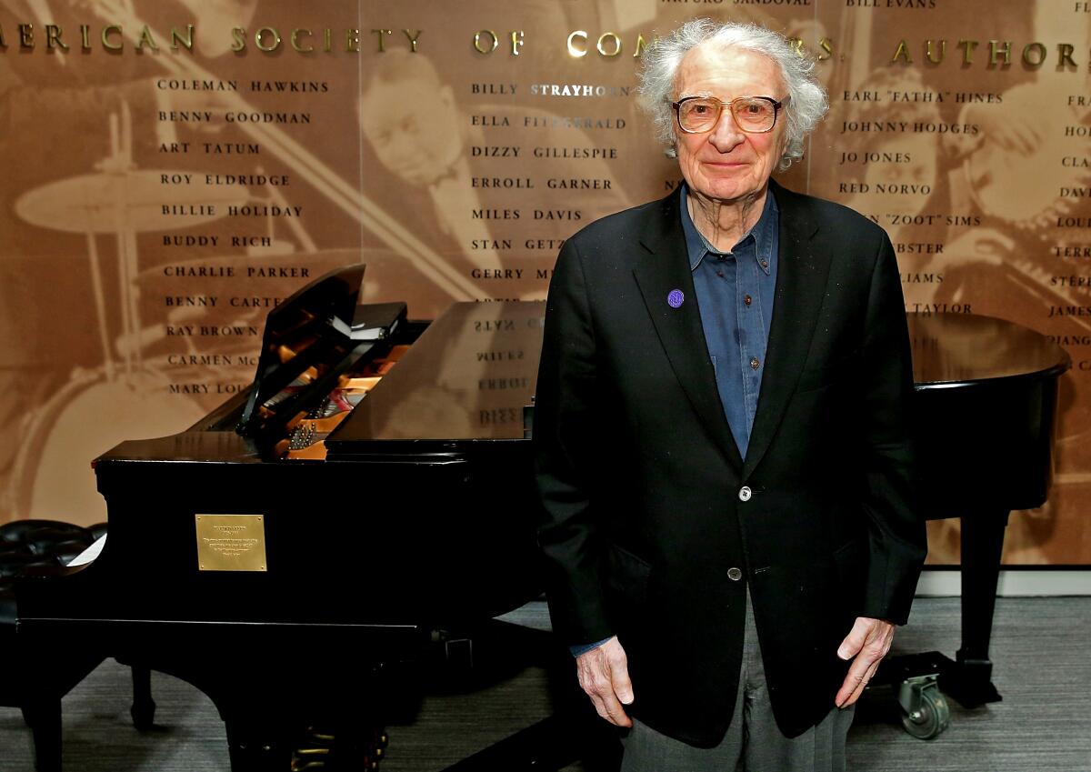 An elderly man stands in front of a grand piano, smiling.
