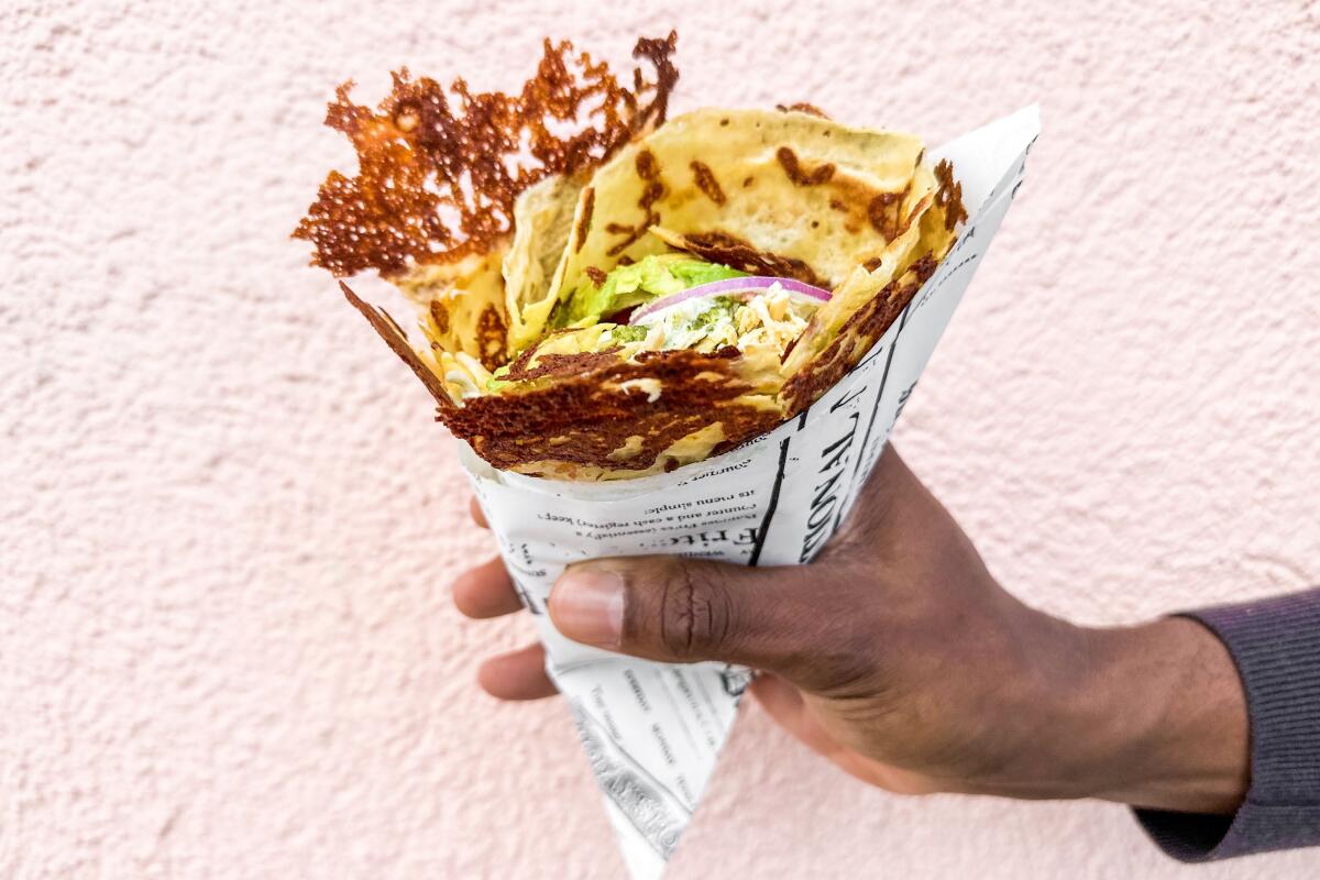 A hand holds a chicken pesto crepe with avocado in a paper cone