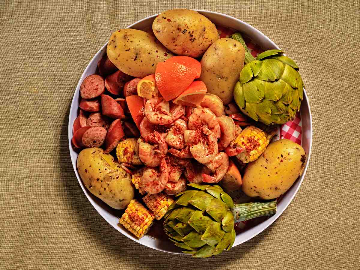 A plate heaped with shrimp, small potatoes, sliced sausage, artichokes, hunks of corn, sections of orange and shrimp.