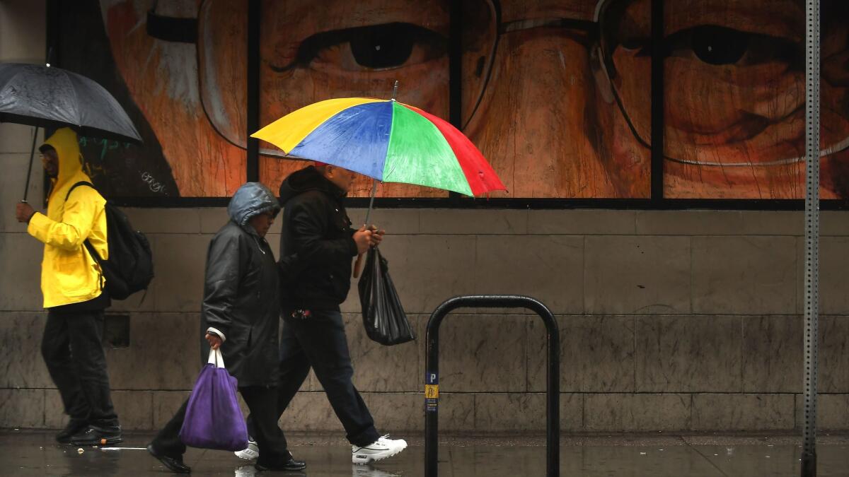 People carrying umbrellas walk past a mural of a close-up of a man wearing glasses on 5th Street.