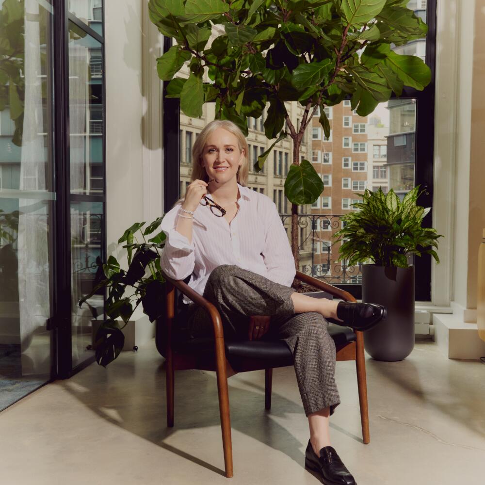 A blond woman, holding eyeglasses in her right hand, sitting in a chair, surrounded by a group of plants.