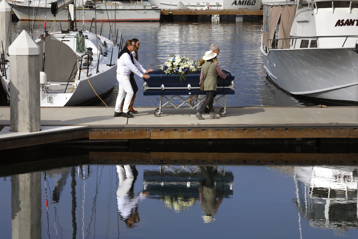 Four people wheel a coffin covered in flowers down a dock toward a boat.