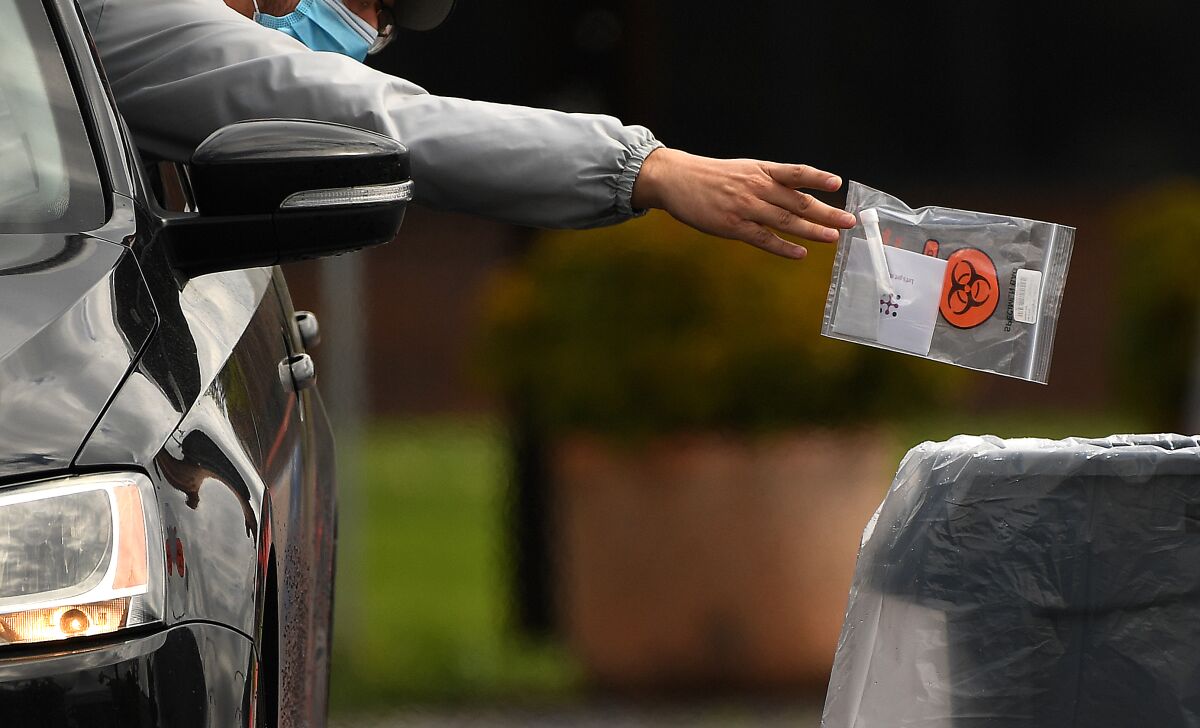 A driver leans out of their window with arm outstretched to drop a bagged coronavirus test specimen into a receptacle