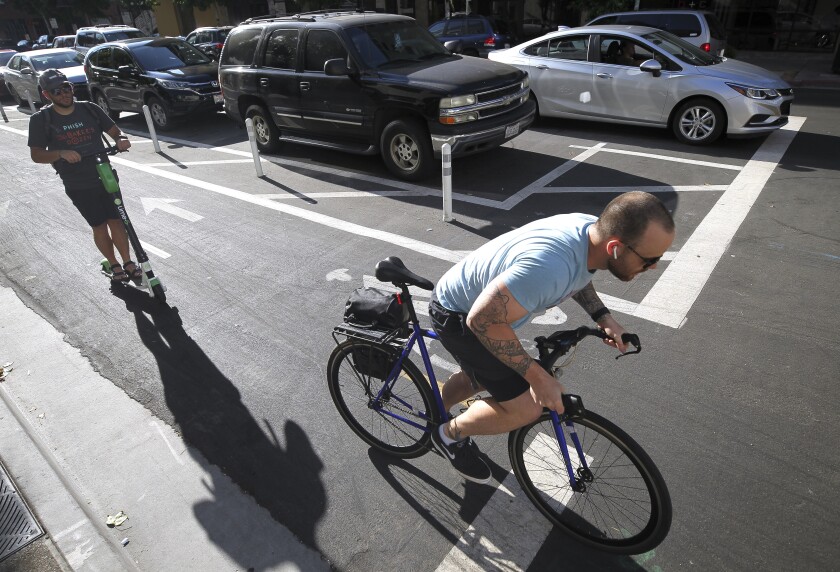 A cyclist and a scooter rider use a bike lane next to where cars park away from the curb, background, on J Street, at the Eleventh Avenue intersection in downtown on Tuesday, July 9, 2019 in San Diego, California.