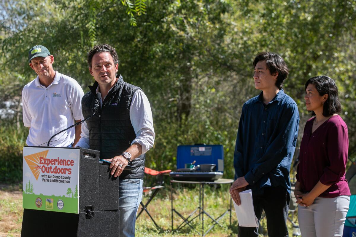 A man stands outdoors at a podium that says "Experience the Outdoors," with three people and vegetation behind him.