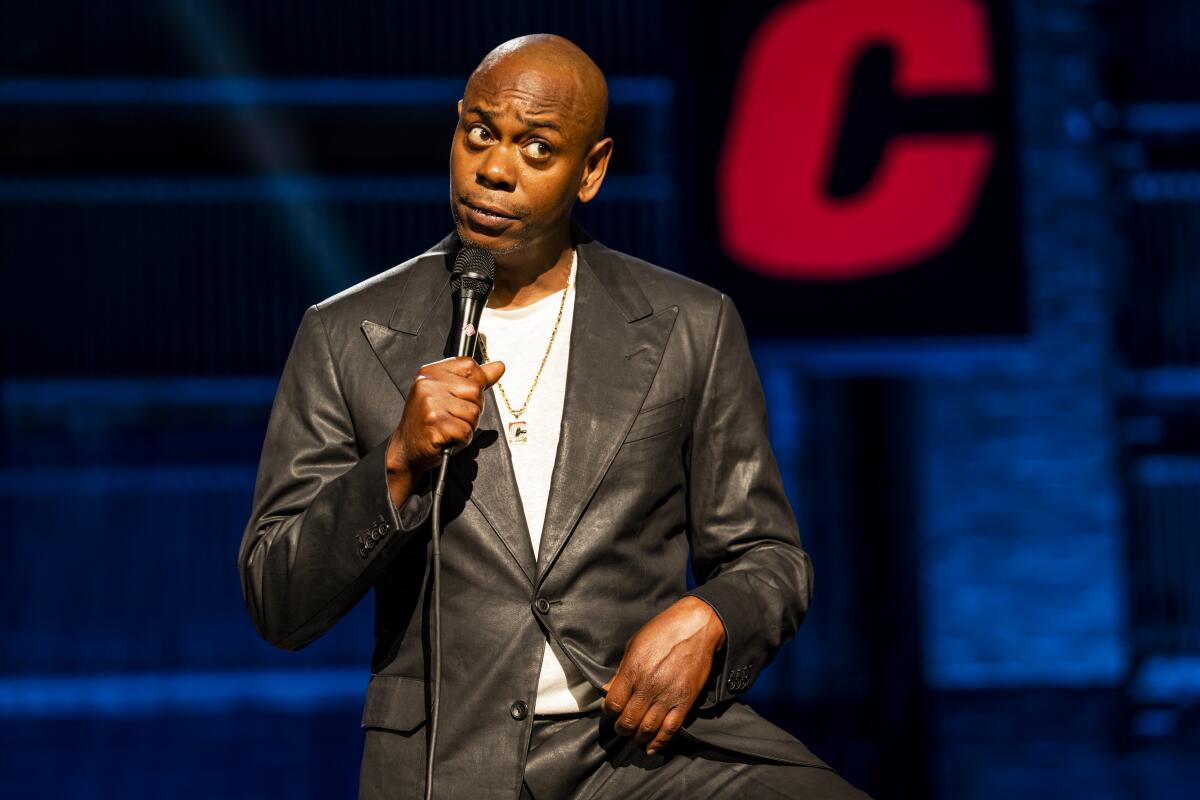 Dave Chappelle in a black leather suit holding a microphone