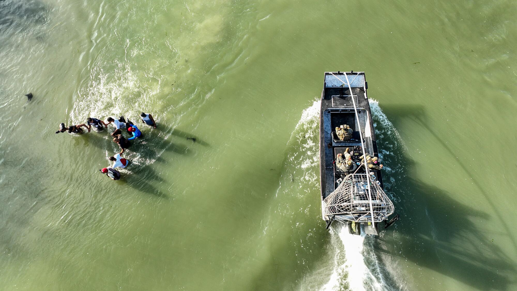 A boat seen from above passes by a group of people wading a river.