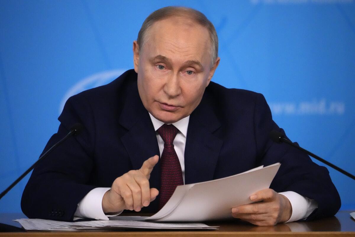 Russian President Vladimir Putin pointing as he holds papers while sitting at a table and speaking into microphones