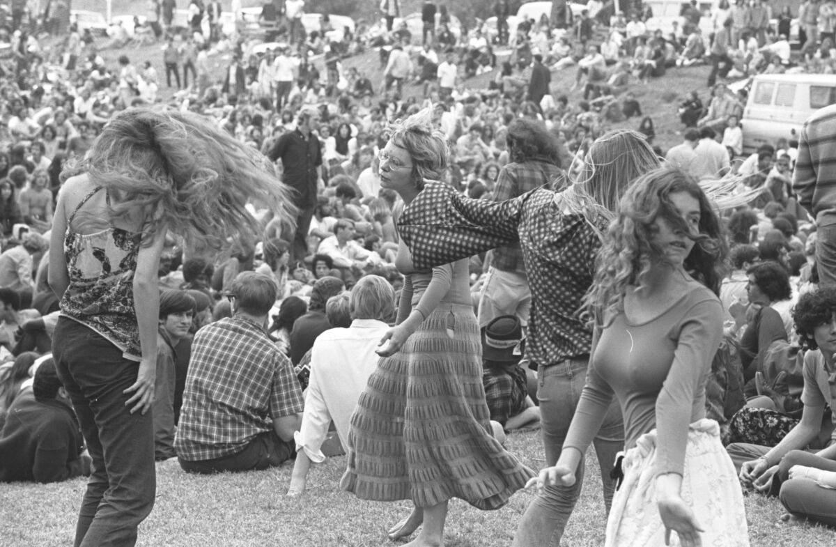 Next to a crowd of people of people lounging on a grassy hill, four women dance.